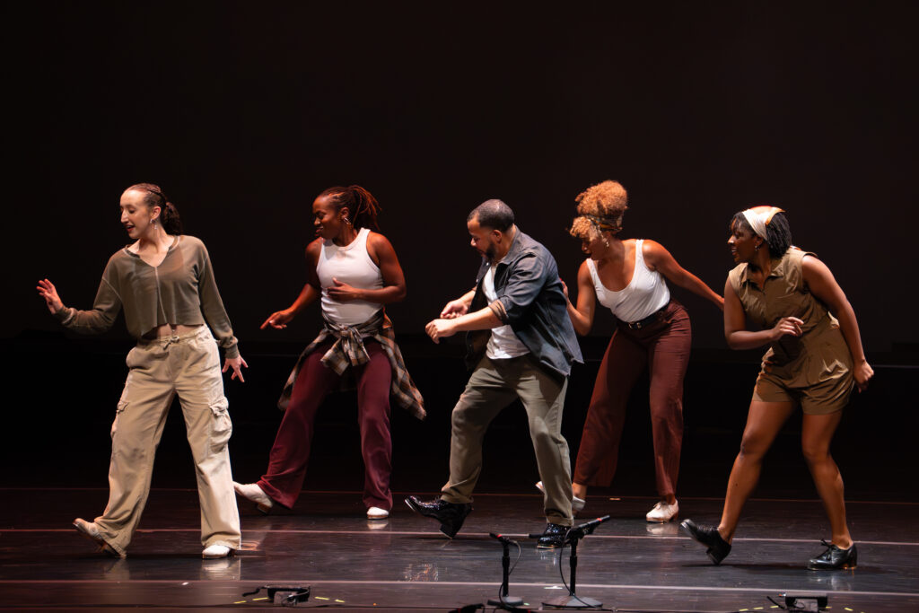 Five percussive dancers wearing tap shoes in mid-performance on a black stage, with instructor and choreographer Ryan K. Johnson in the middle.