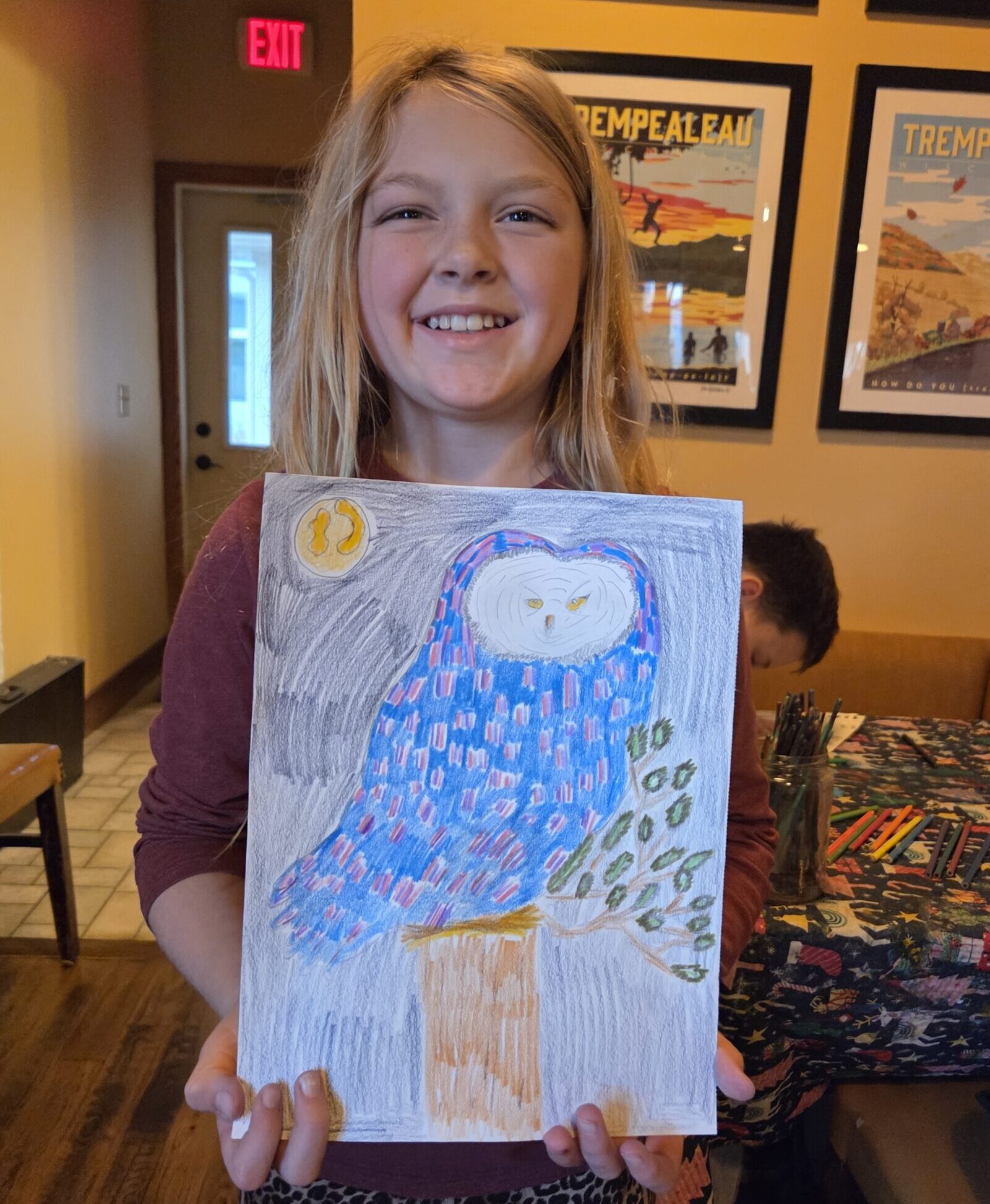 A girl with blonde hair and light skin smiles and holds a drawing of an owl.