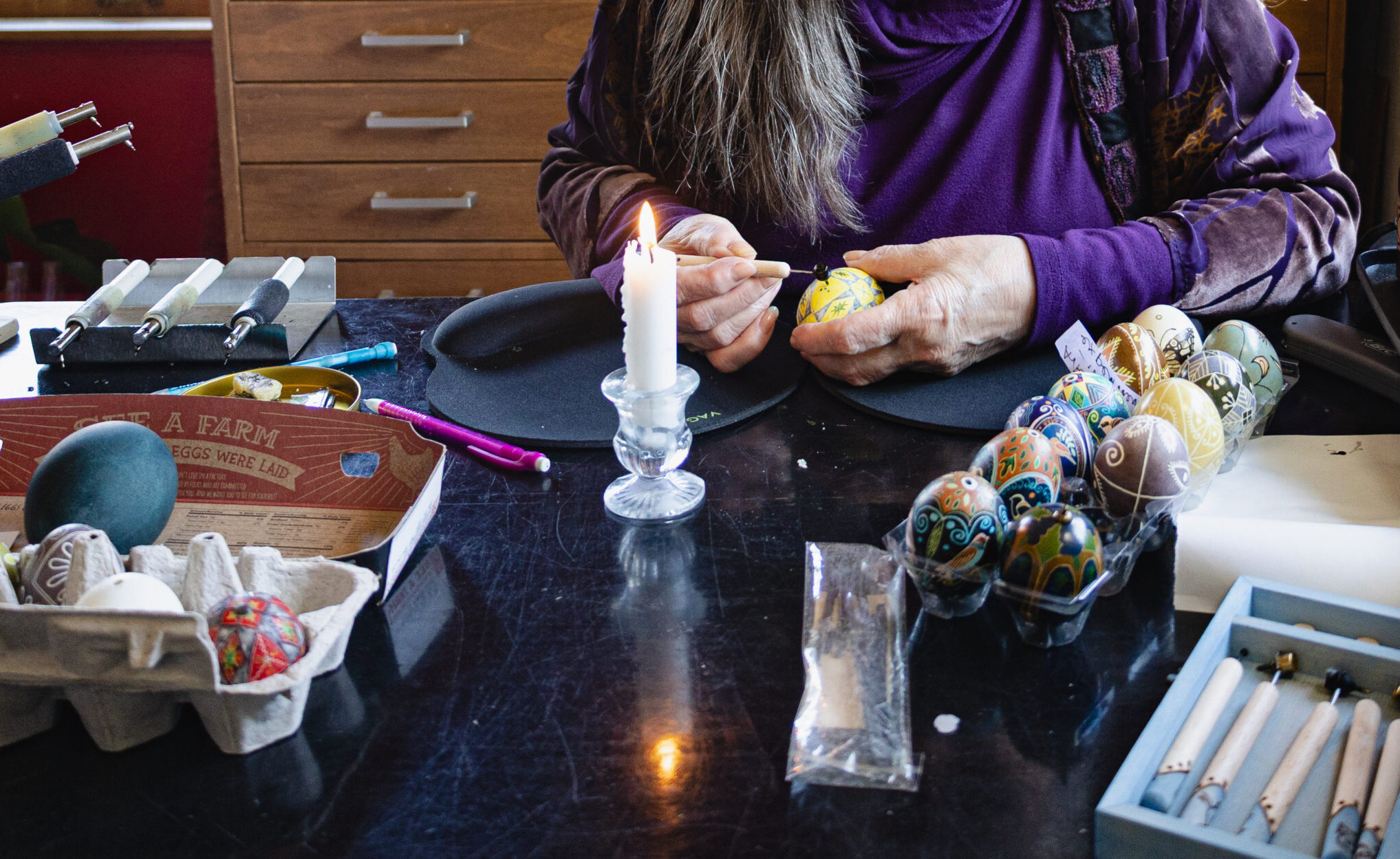 A person sitting at a table applying wax to a painted egg. There are other colorful eggs by them in a tray and a lit candle as well as other tools.