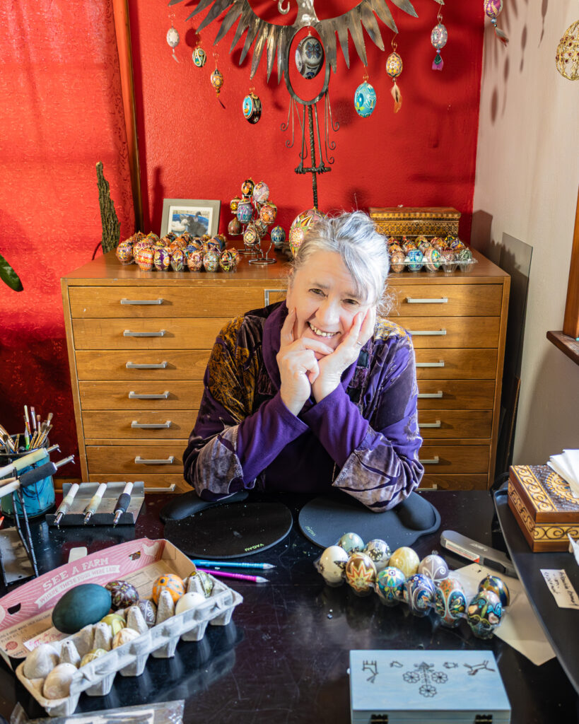 A person sitting at a table and smiling. They have painted eggs in front of them and other tools.