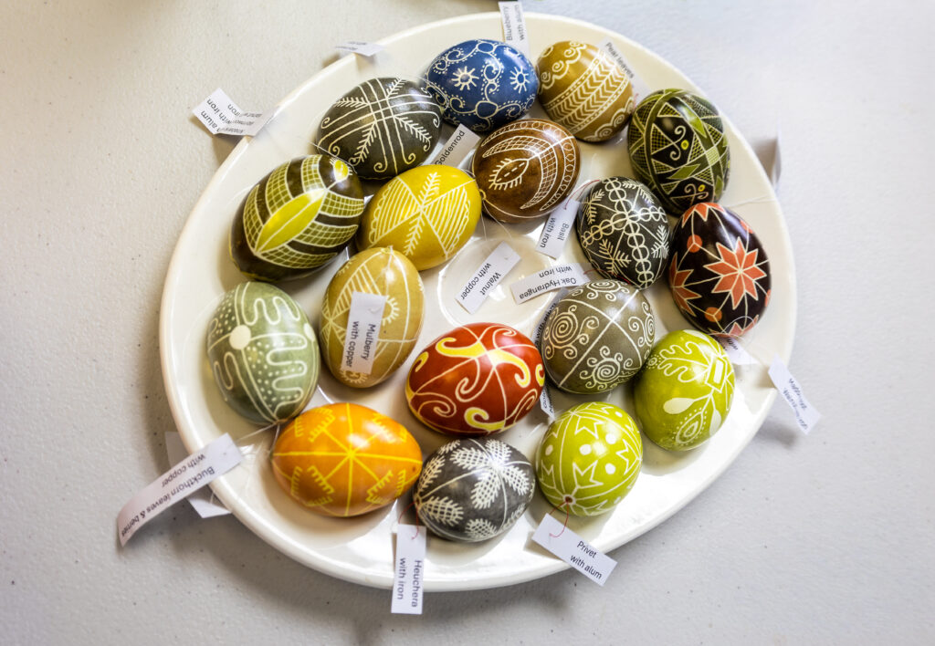 A plate of brightly colored eggs decorated with intricate wax-resist patterns, each labeled with the natural dye used, including onion skin, indigo, and other plant-based materials.