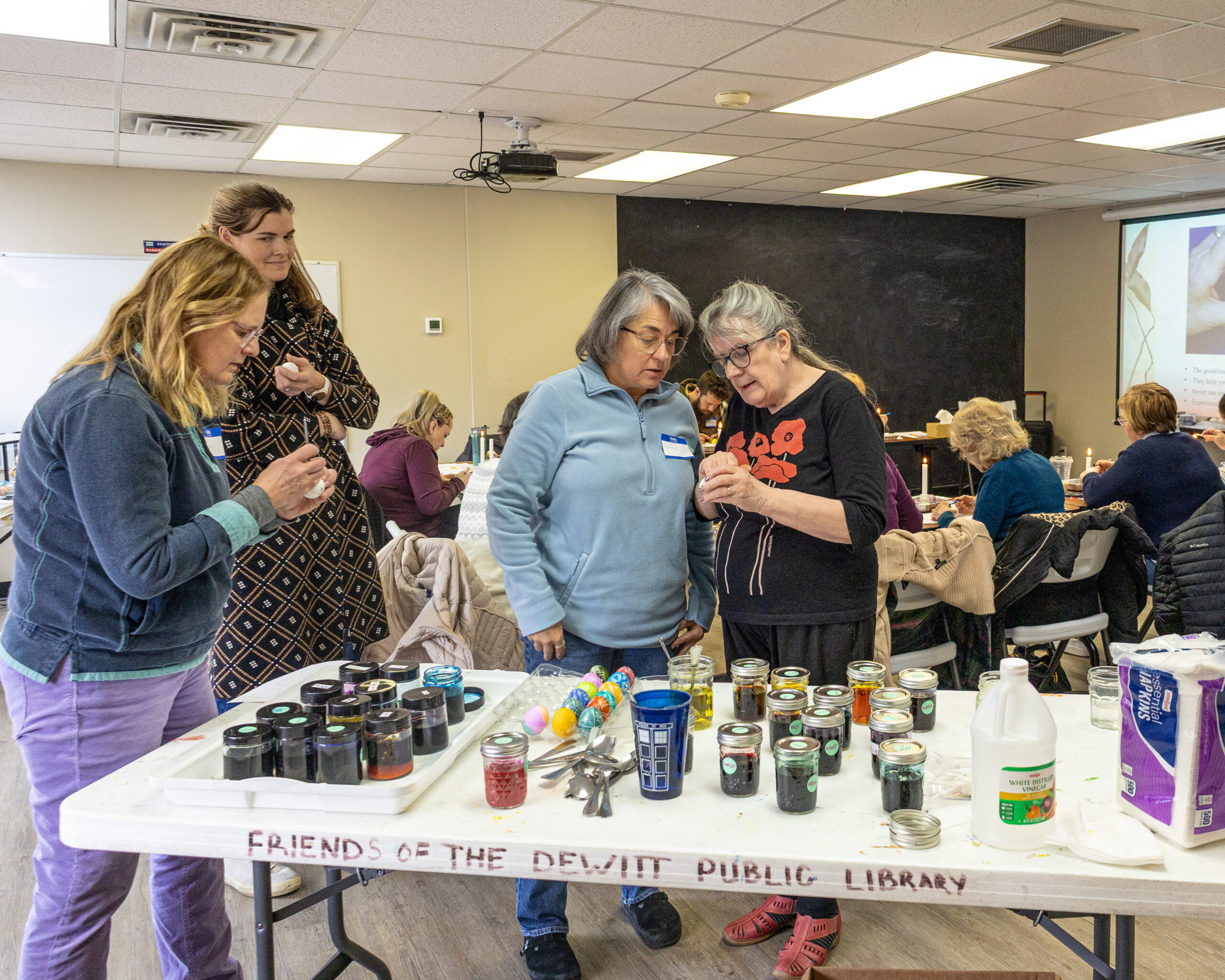 A person standing next to three others as they look on and listen to instructions during an egg painting workshop.
