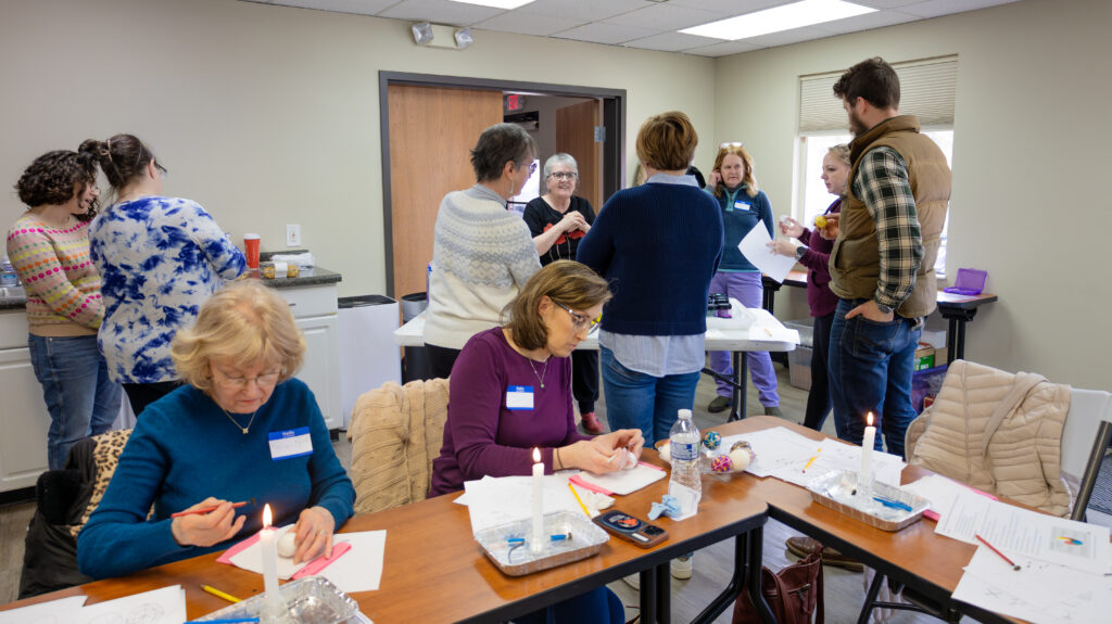 A group of people standing around a table as they talk to an instructor. Two others sit at a table holding tools by lit candles as they work on applying wax to eggs.