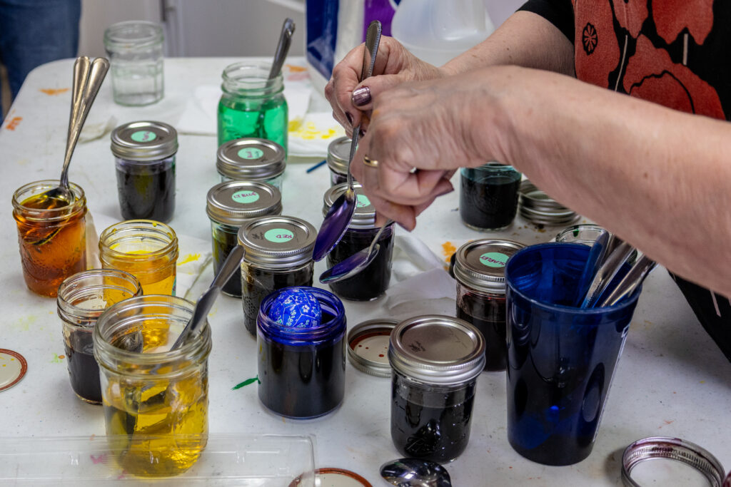 A person using two spoons to rotate a painted egg that has been dipped in a blue liquid dye. There are other jars on the table with dark and yellowy liquids.