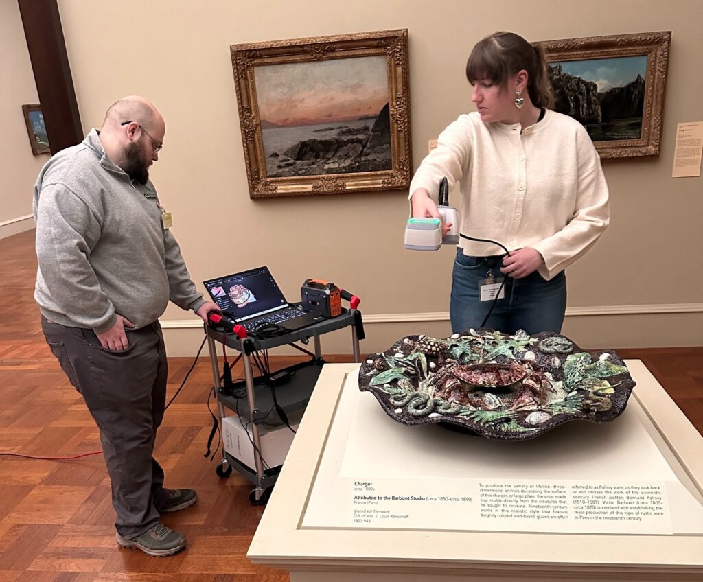 Two people analyzing a ceramic artwork in a museum using a scanning device. A laptop on a cart shows digital results. Paintings hang in the background.