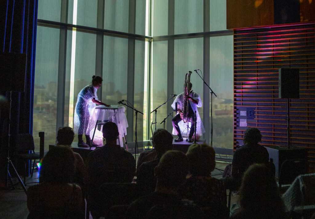 Two people wearing bubble wrap playing instruments on a dark stage.
