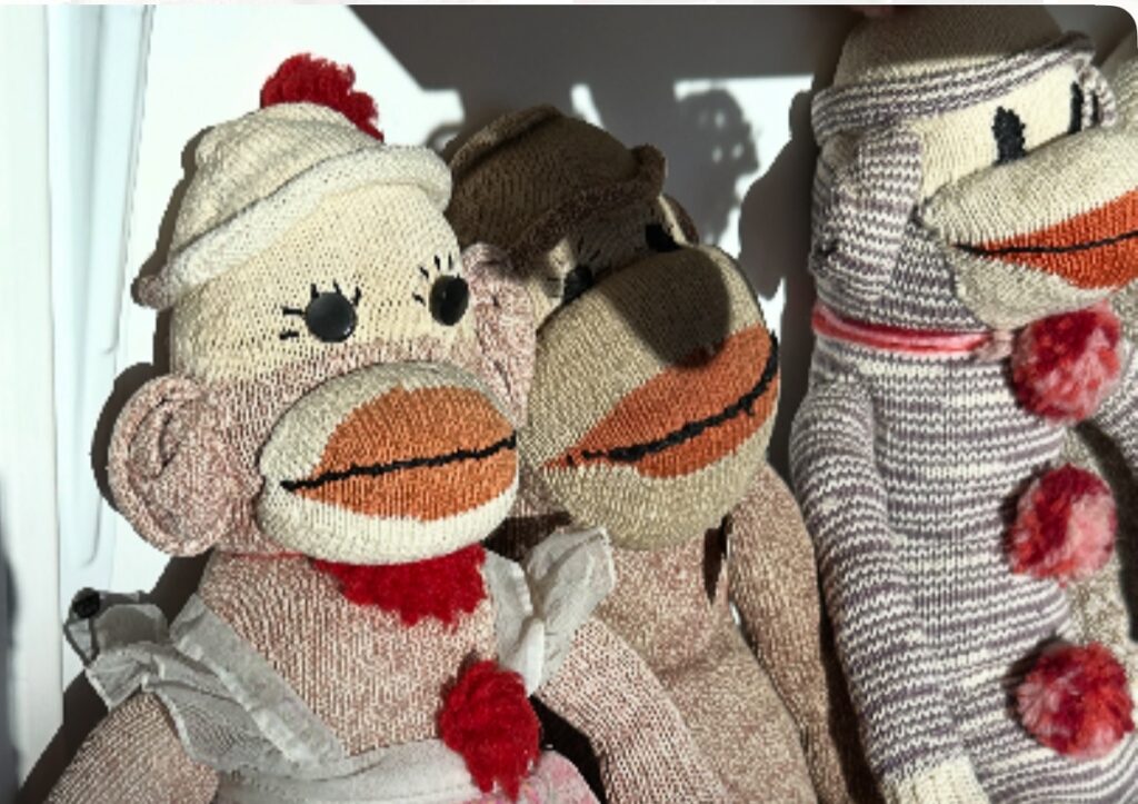Three sock monkey dolls with vibrant expressions, adorned in varied hats, pom poms, and scarves.