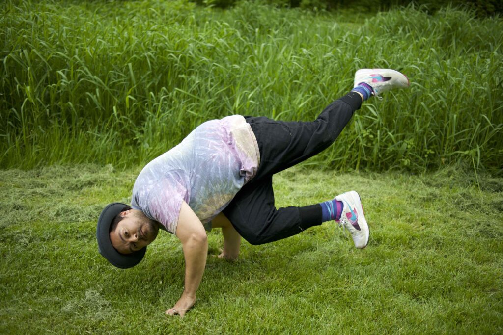 A man wearing a tie-dyed shirt, a wide-rimmed hat, colorful socks, and sneakers breakdancing outdoors on a green lawn.