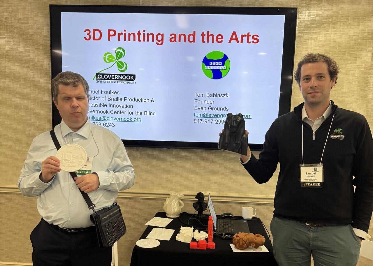 Two men standing in front of a screen displaying "3D Printing and the Arts." One holds a 3D-printed disc, the other a 3D printed bear form. There is a table with 3D prints between them.