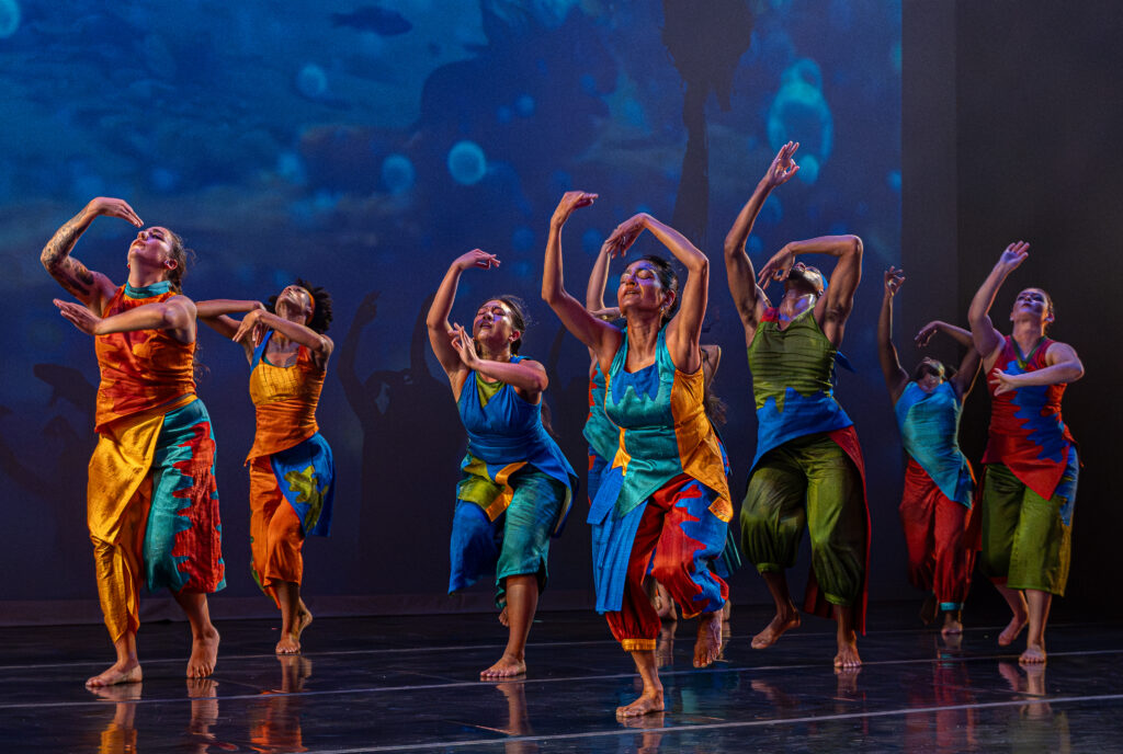 A group of seven barefoot dancers perform on a lit stage in vibrant, colorful draped costumes of orange, teal, blue, and green. Their arms are raised and faces tilted upward, with a blue ocean-like projection behind them.