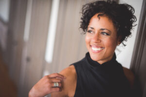 Headshot of a smiling woman with short, brown curly hair, medium skin town, wearing a black shirt.