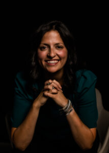 A smiling, dark-haired, olive-skinned woman leaning forward with her hands clasped in front of her, wearing a dark green top and a stack of bracelets.