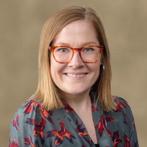 Headshot of a woman with blonde hair, light skin, brown glasses, and a navy blue floral shirt.
