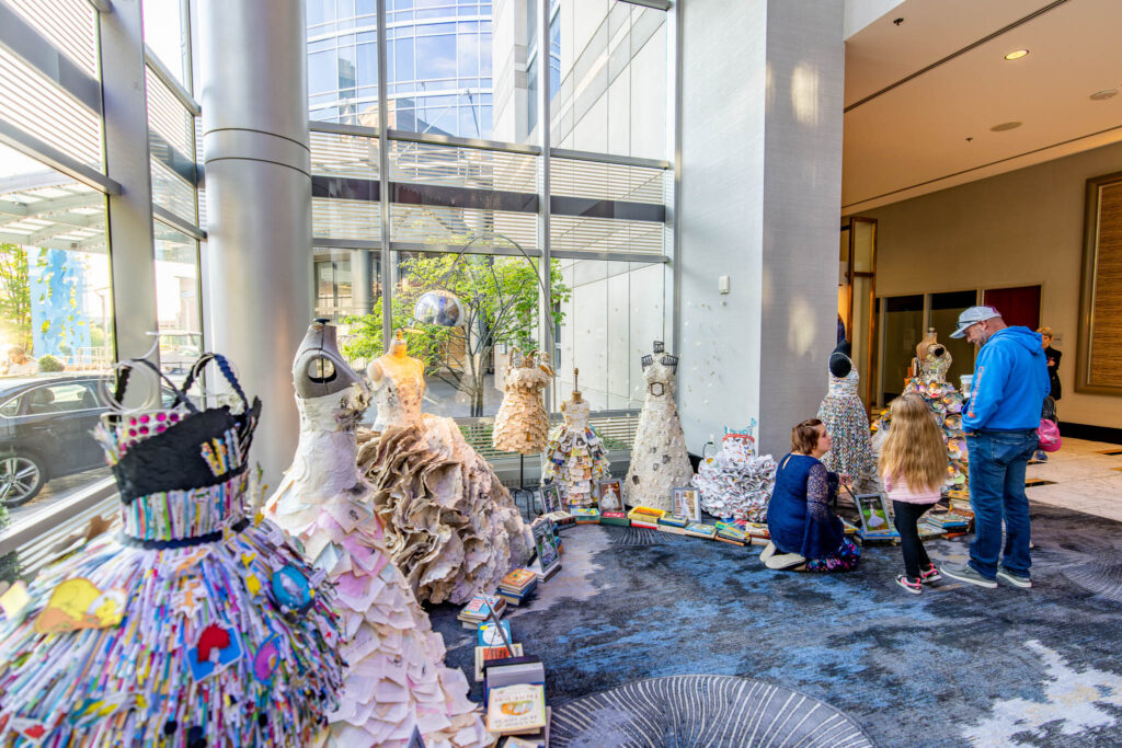 Three people looking at colorful dresses and gowns displayed by large windows.