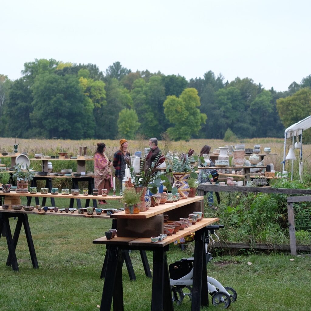 People browse and chat in an outdoor pottery market with wooden tables displaying colorful ceramics around a lush green field with a forest in the background.