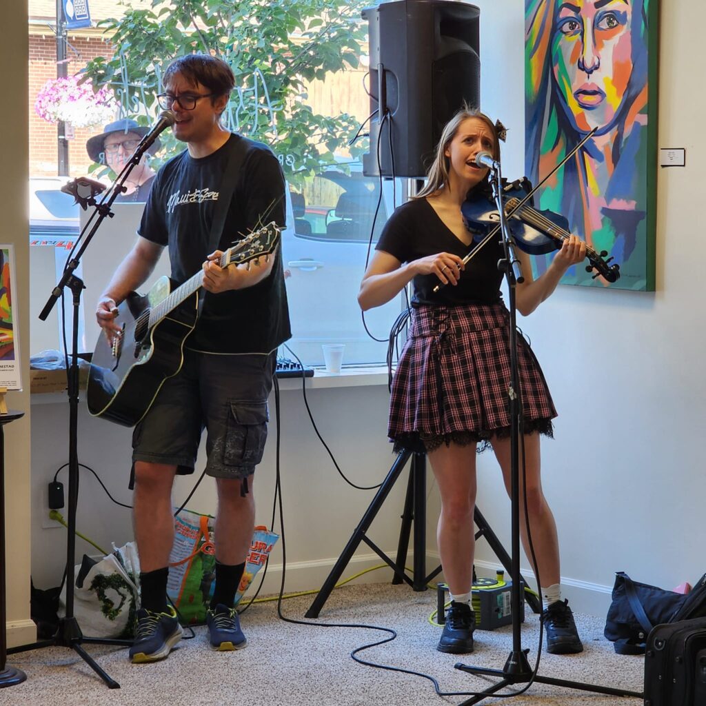 Two musicians - one playing acoustic guitar, the other playing a blue electric violin - singing into microphones inside near a window. A brightly colored painting of a woman hangs on the wall behind the violinist.