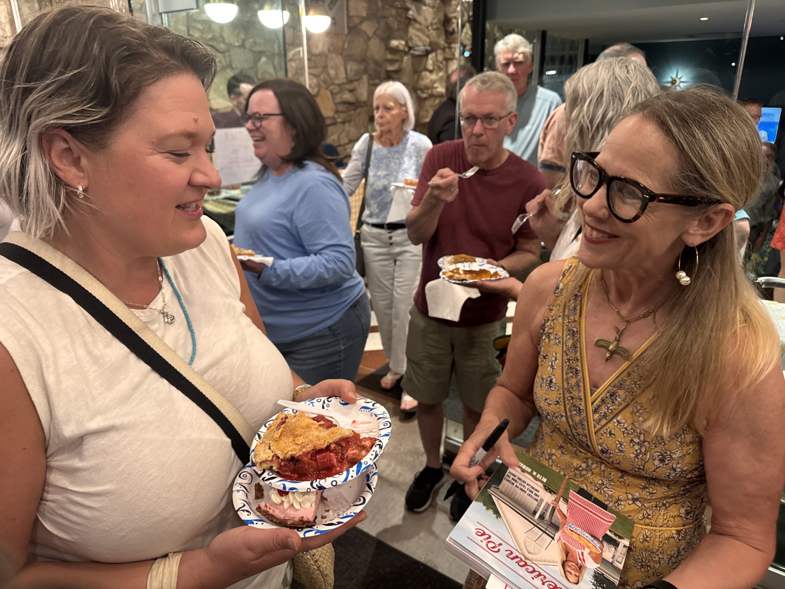 A smiling woman in a yellow dress signs books while another person holds two plates of fruit pie at a crowded event.