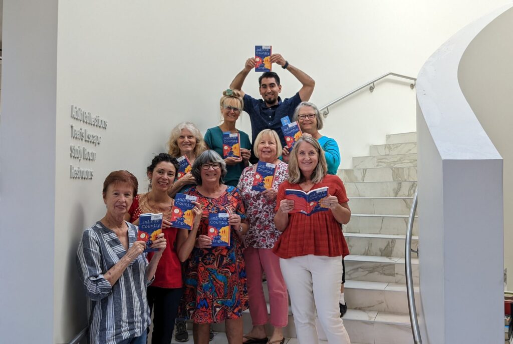 A group of nine people stands on a spiral staircase, each holding up the same book, "Infinite Country" and smiling.