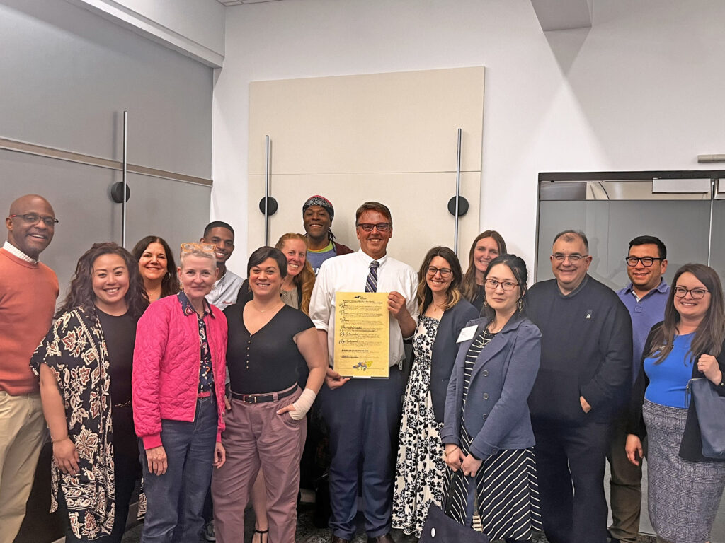 A group of Arts Midwest staff and Board members gather in a conference room, smiling as they stand around Brian Tiemann (center). Brian holds a framed notice designating an honorary Board seat in his name, recognizing his leadership and service