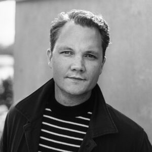 Black and white photo of Carl, looking right at the camera with wavy hair, a striped sweater, and dark jacket.