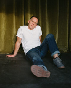 Casual day lit portrait of the artist in his studio. Light skin tone, with short dark hair in a reclined posture dressed in a white t-shirt and blue jeans with sneakers. The angle of the photo shows the bottom of the shoes with a lush green velvet backdrop hanging in the background.