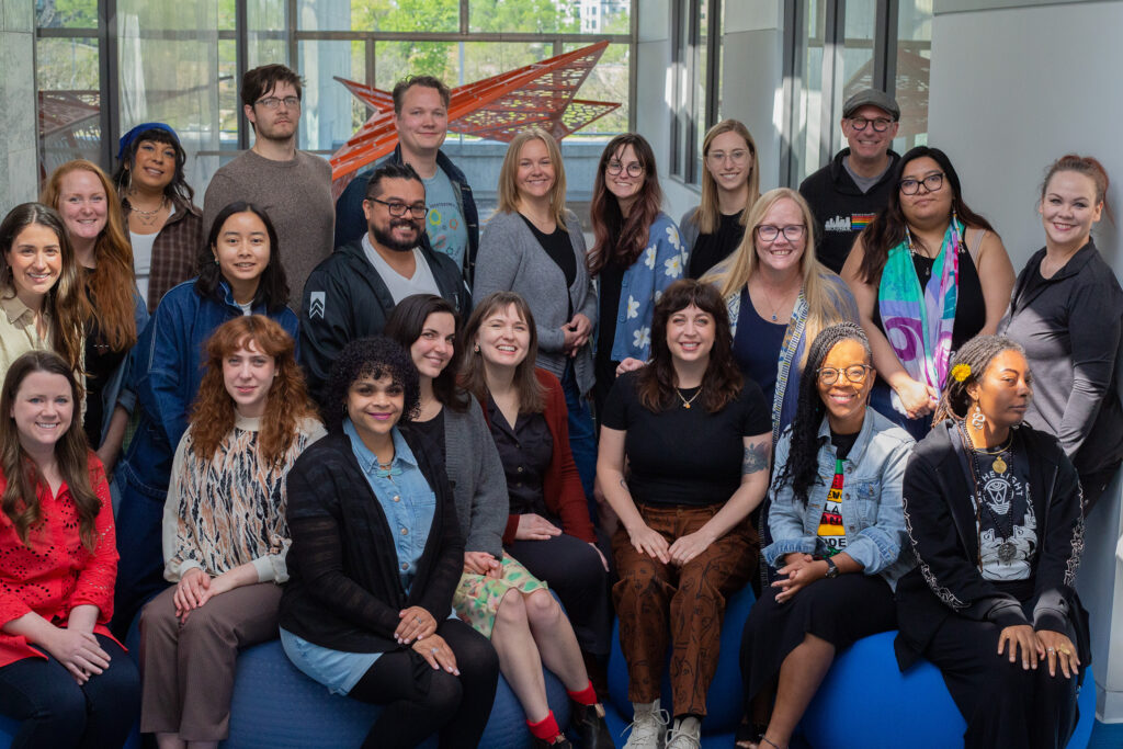 A diverse, smiling group of 22 people poses indoors near large windows, with a modern artwork in the background.