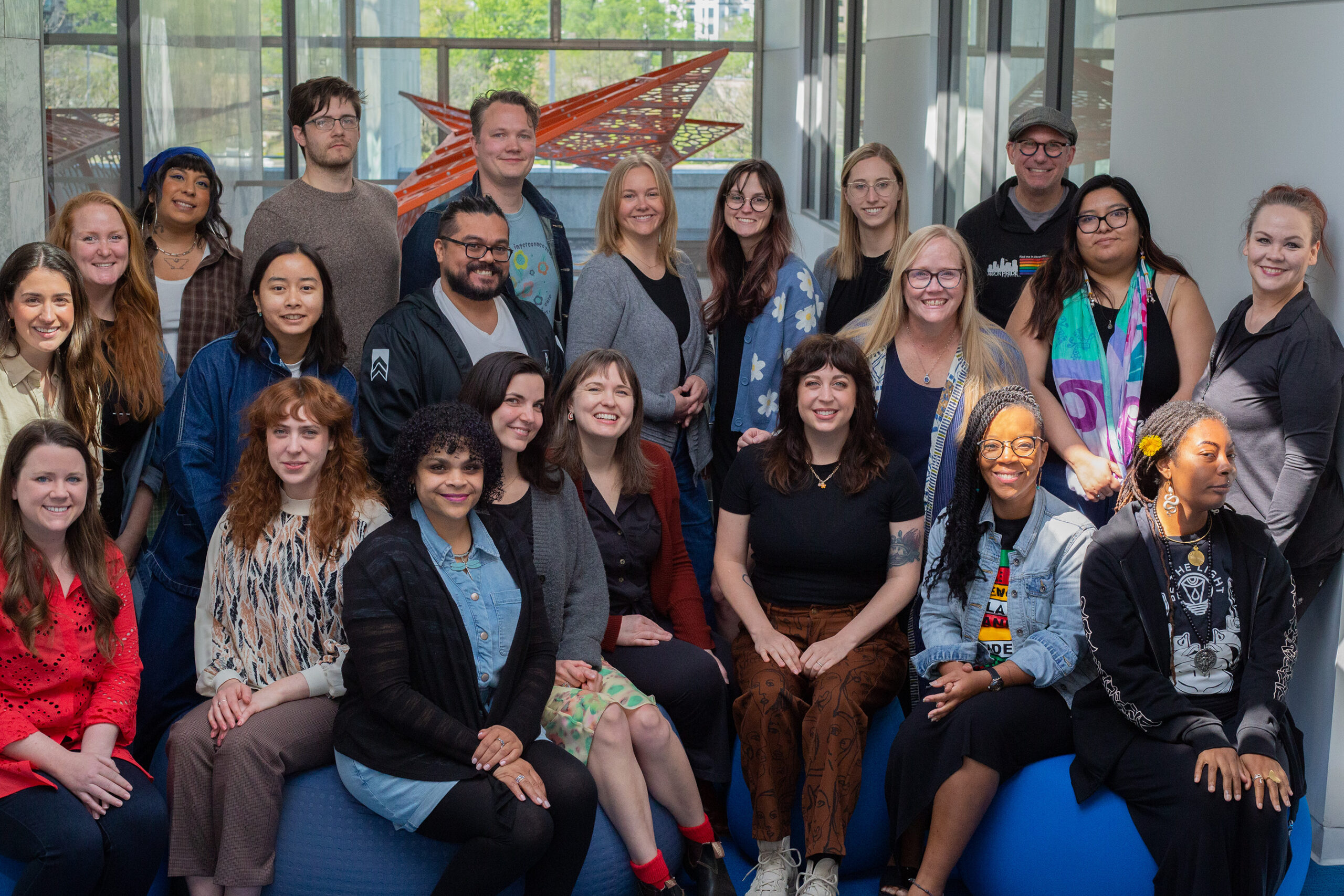A diverse, smiling group of 22 people poses indoors near large windows, with a modern artwork in the background.