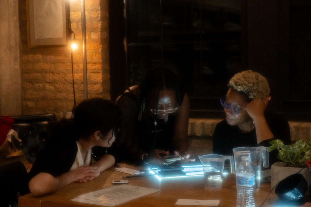Three people with dark skin sitting at tables with film photography equipment in a dimly lit room.