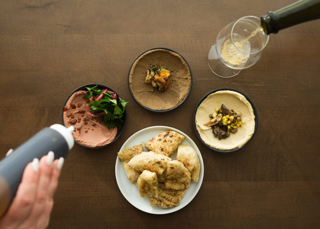 Three round bowls filled with colorful spreads topped with vegetables, plus a fourth round bowl filled with chunks of bread. A hand holding a squeeze bottle hovers over one of the bowls. A bottle of wine is poured into a glass net to the top bowl.