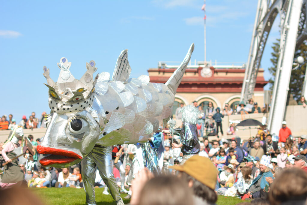 A whimsical parade scene with a person in a large, metallic silver fish costume adorned with a crown, surrounded by a festive crowd. Bright, sunny sky and an arched building in the background convey a joyful, celebratory atmosphere.
