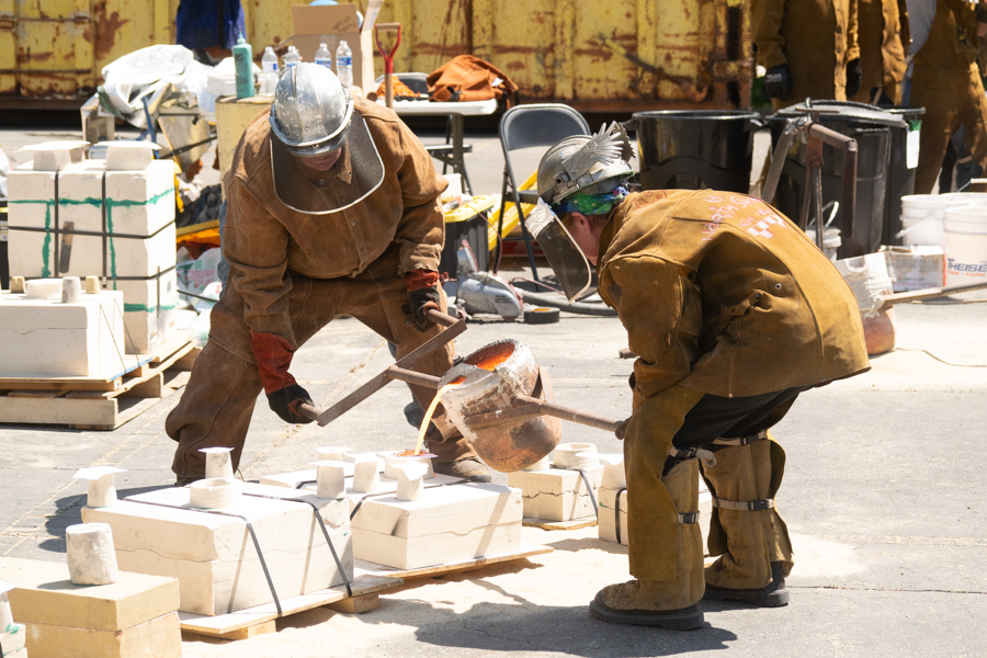 Two people in heavy iron casting protective gear pouring hot metal into molds that are resting on the ground.