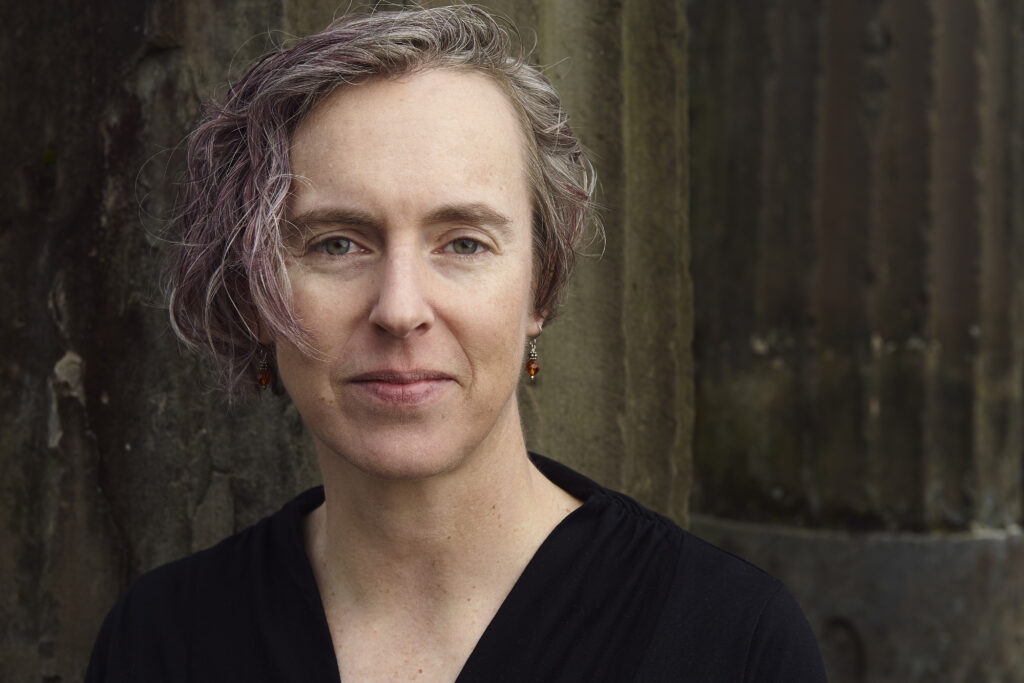 A person of light skin tone with short hair smiles at the camera. Their hair is grey with streaks of pinkish purple.