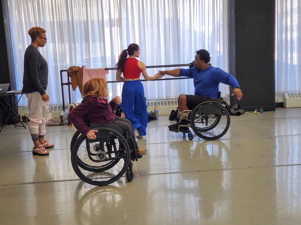 A dance class setting with four dancers. One dancer in a wheelchair is holding hands with a standing dancer as they both move. Beside them are two other dancers, one in a wheelchair, one standing, watching their movements.