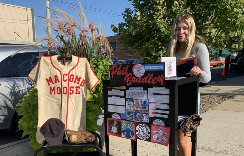 A young person standing outdoors by a table with a display of information and a historic baseball jersey.