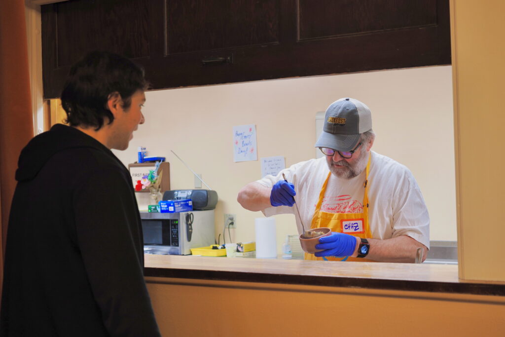 A man wearing a baseball cap and blue gloves serving up soup into a small bowl behind a counter separating him from another person/