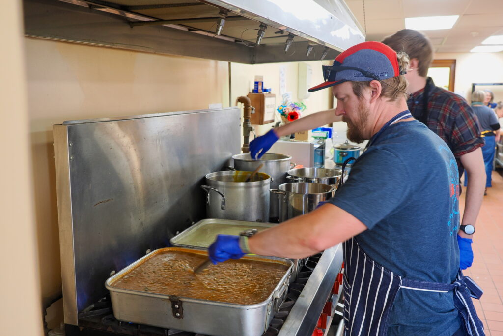 Two pep=ople with light skin wearing blue gloves stir large pans and pots of soup.