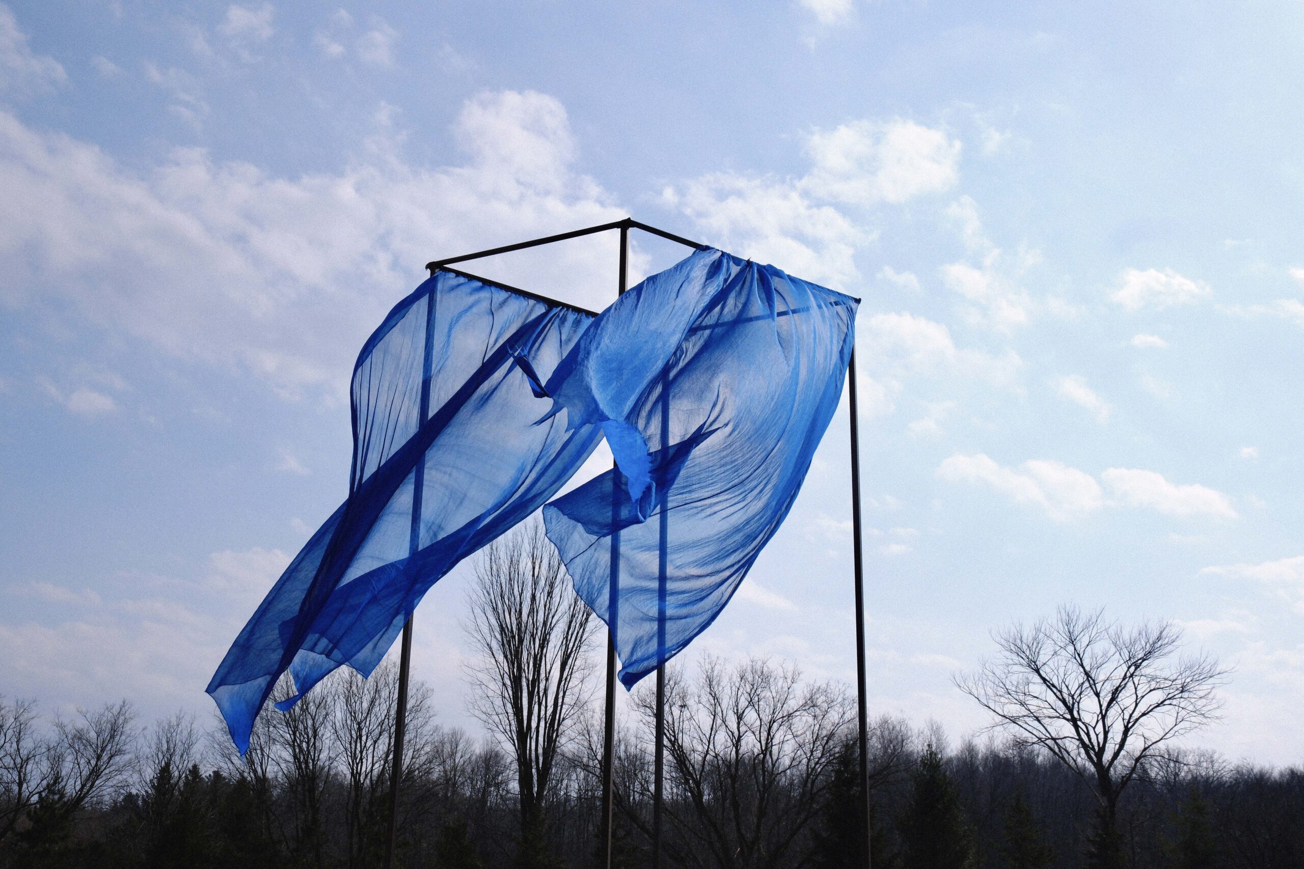 A large, translucent blue fabric billows in the wind, draped over a tall black metal frame outdoors. Bare trees line the horizon beneath a pale blue sky with scattered clouds, emphasizing the fabric’s movement and lightness against the winter landscape.