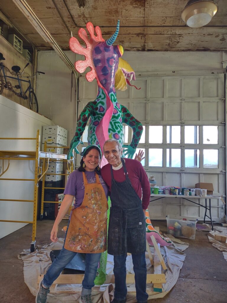 Two people standing next to each other with a large colorful sculptural work behind them