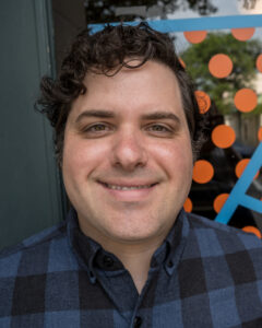 A person of medium skin with short curly hair smiles at the camera.