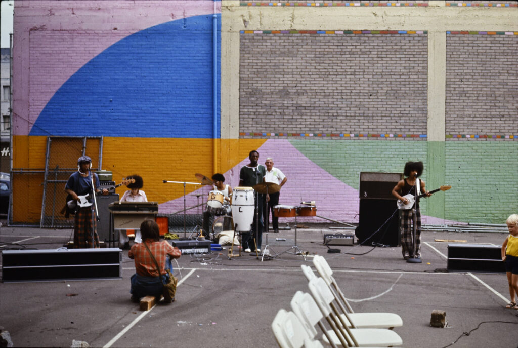 An archival photo showing a band performing outdoors. They are standing by a side of a building with a colorful mural