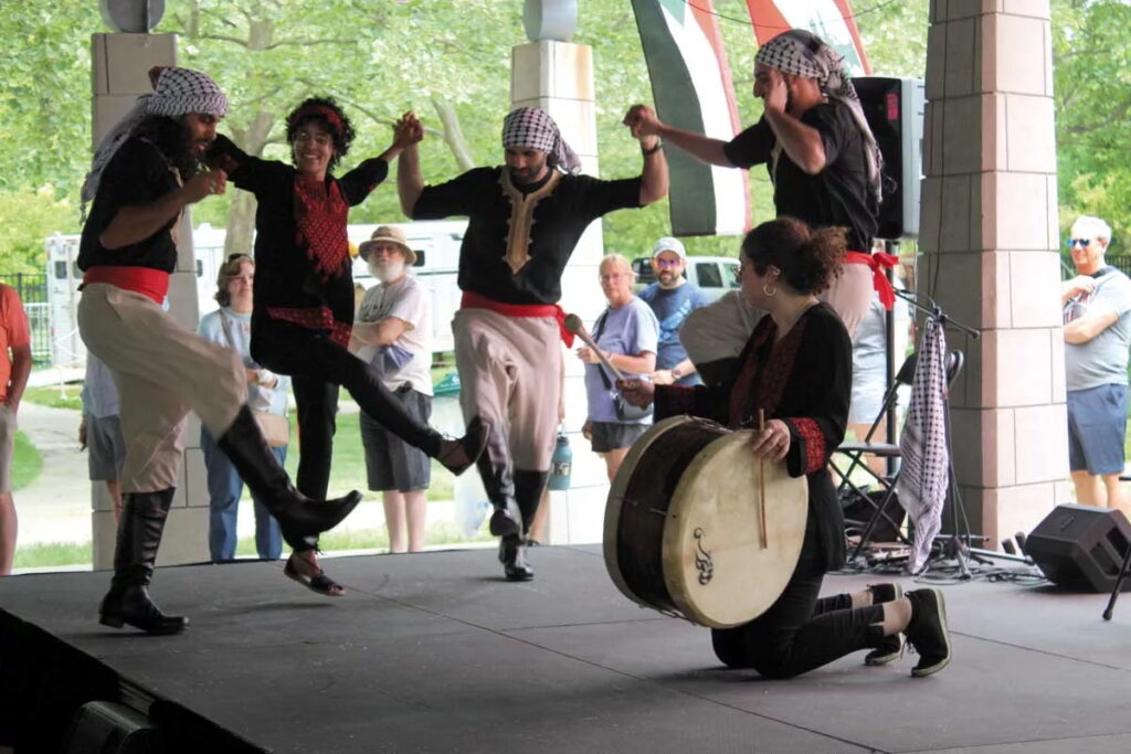 Four people holding hands and dancing while another person kneels and bangs a drum.