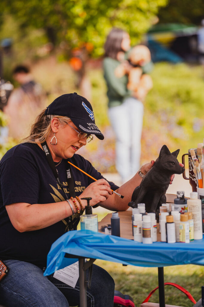 A woman with light skin, a blonde ponytail, and a black Veteran cap painting a small sculpture of a wolf at an outdoor table.