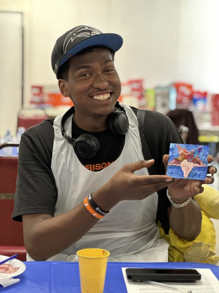 A veteran with dark skin, a dark baseball cap, and a plastic apron smiling and holding up a small painting of a star.