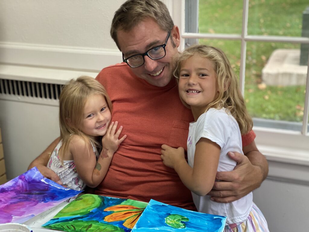 A veteran with glasses and short blonde hair with his arms around two young girls with blonde hair, sitting at a table with three colorful paintings in front of them.