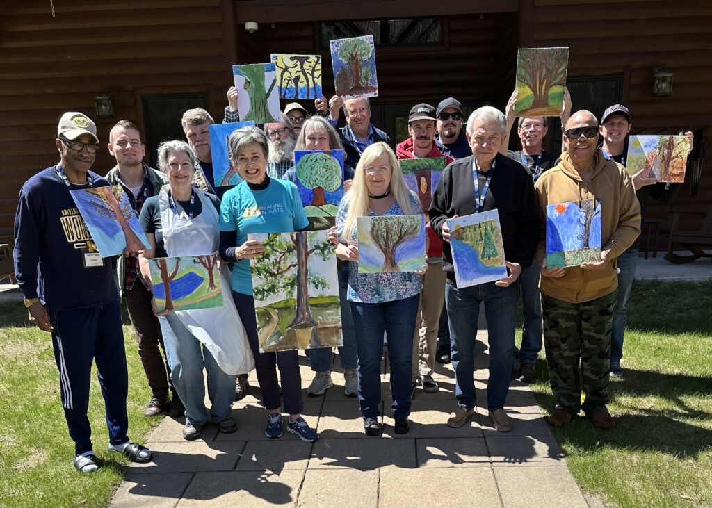 Sixteen veterans of various ages, genders, and backgrounds, holding up their original paintings.