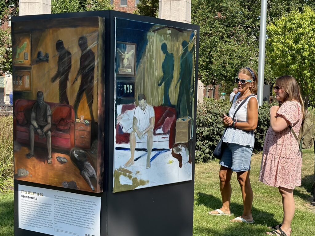 An outdoor art installation featuring two original paintings by a veteran. Two observers stand nearby to take in the art.
