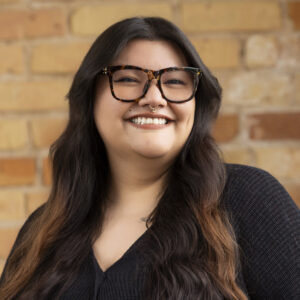Headshot of a smiling person of medium skin tone, with dark brown hair, glasses, and a nose ring, and wearing a black sweater.