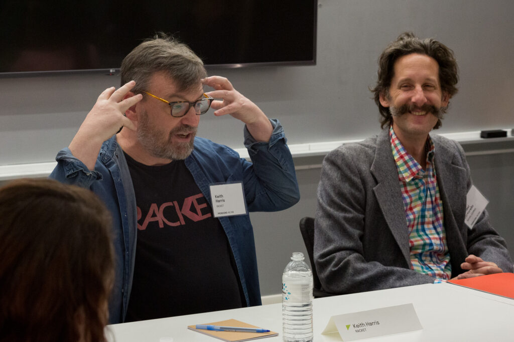 Two people sitting at a table, facing forward. One of them is smiling as they other gestures with their hands close to their head.