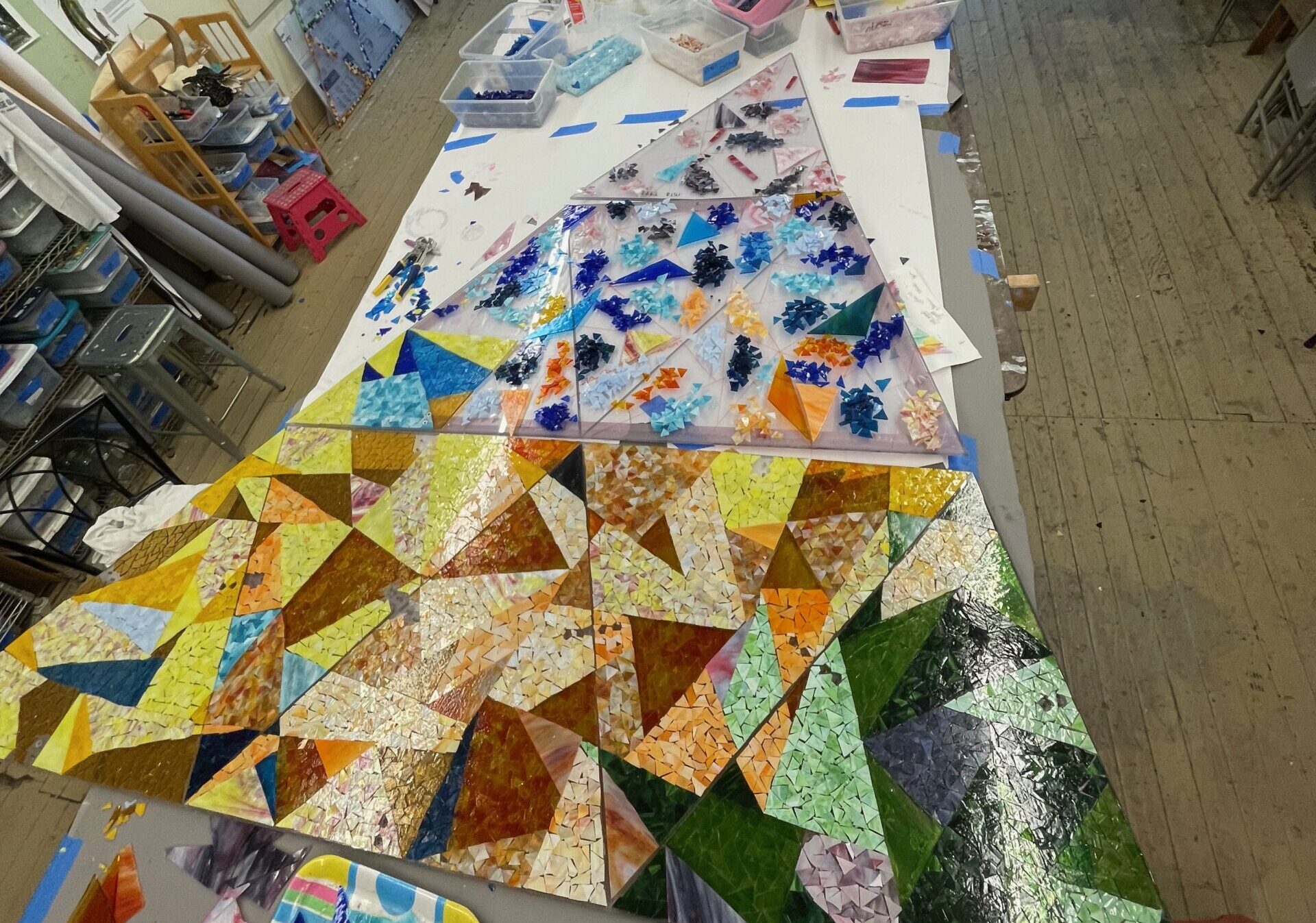An in-progress mosaic of large, colorful glass triangles forming a larger triangle on the floor of an art studio, with paintings and art supplies lining the walls.