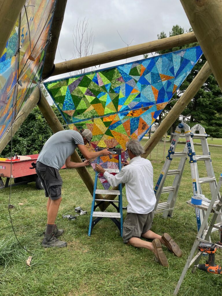 A craftsperson kneeling on green lawn while another stands next to them as they install a panel of colored glass in a large mosaic in the shape of a triangle, forming one side of an open pavilion in a public park.